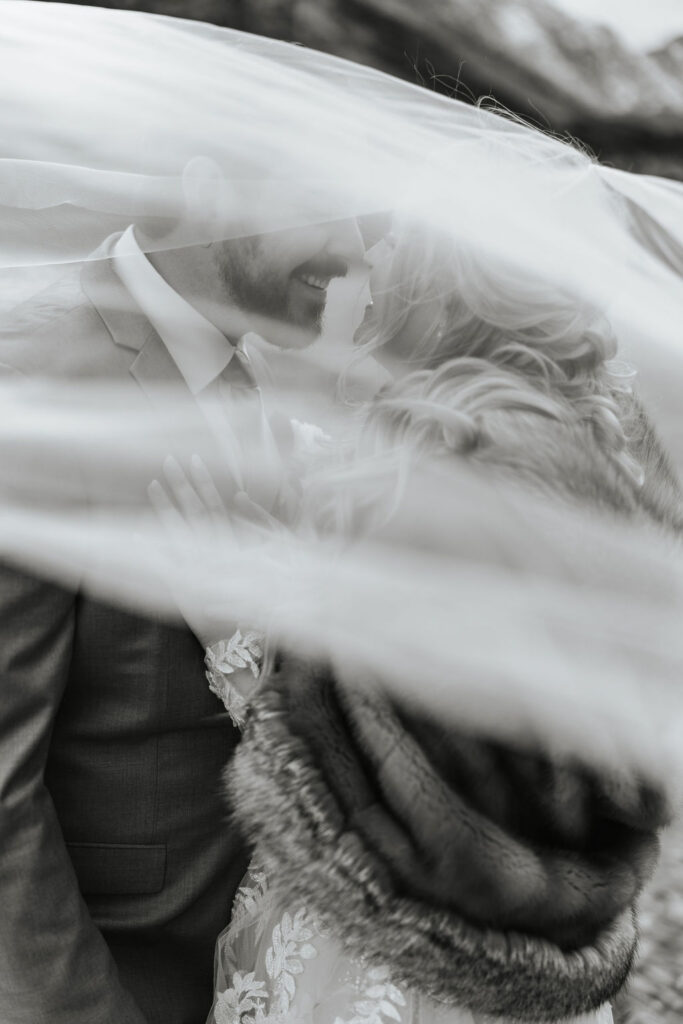A black and white photo of a couple stands in their wedding outfits with a veil around them in the rocky mountains of colorado